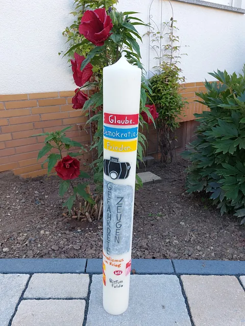 Große, farbig gestaltete Kerze steht vor einem Rosenstrauch