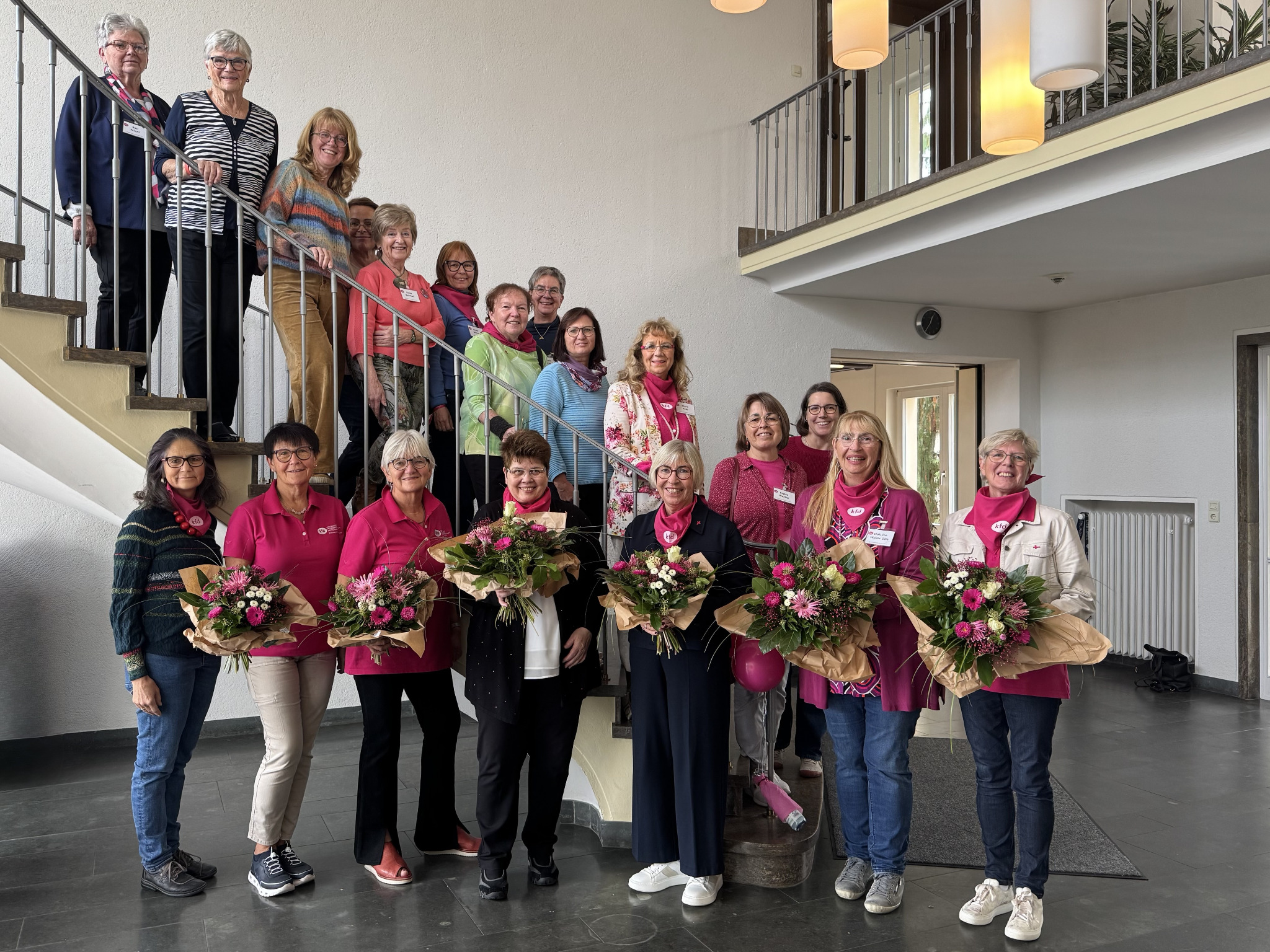 Gruppe von bunt gekleideten Frauen stehen vor und auf der Treppe. 6 Frauen vorne mit Blumenstrauß