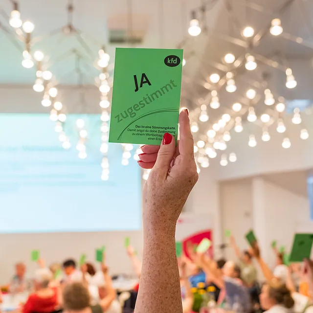 Im Vordergrund eine Frauenhand, die eine Ja-Karte in die Höhe hält