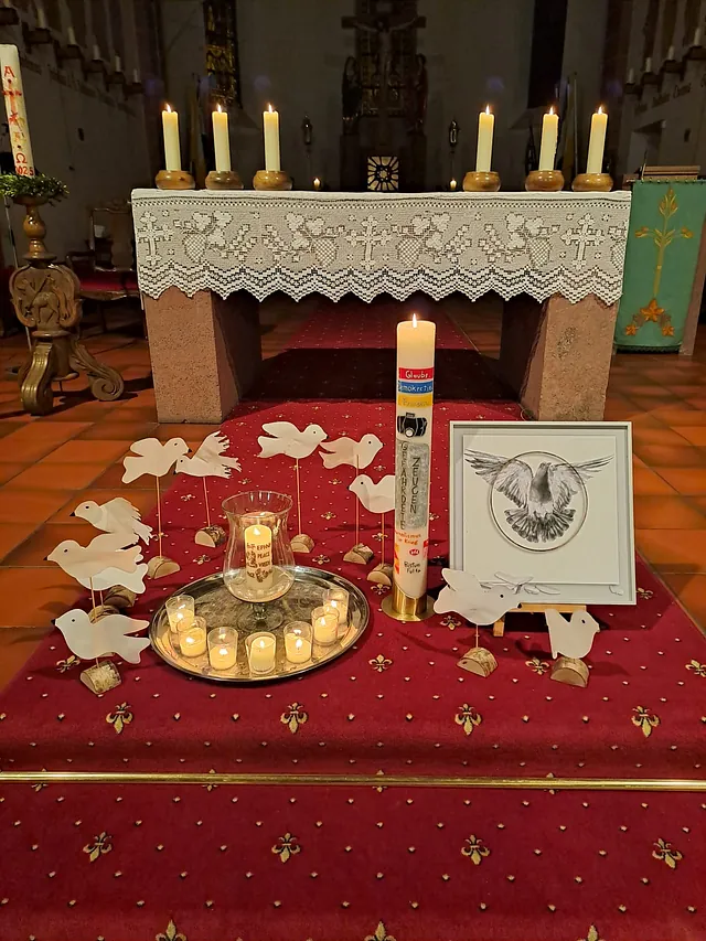 Vor dem Altar stehen viele kleine Kerzen und eine große Kerze, umrahmt von gebastelten weißen Tauben